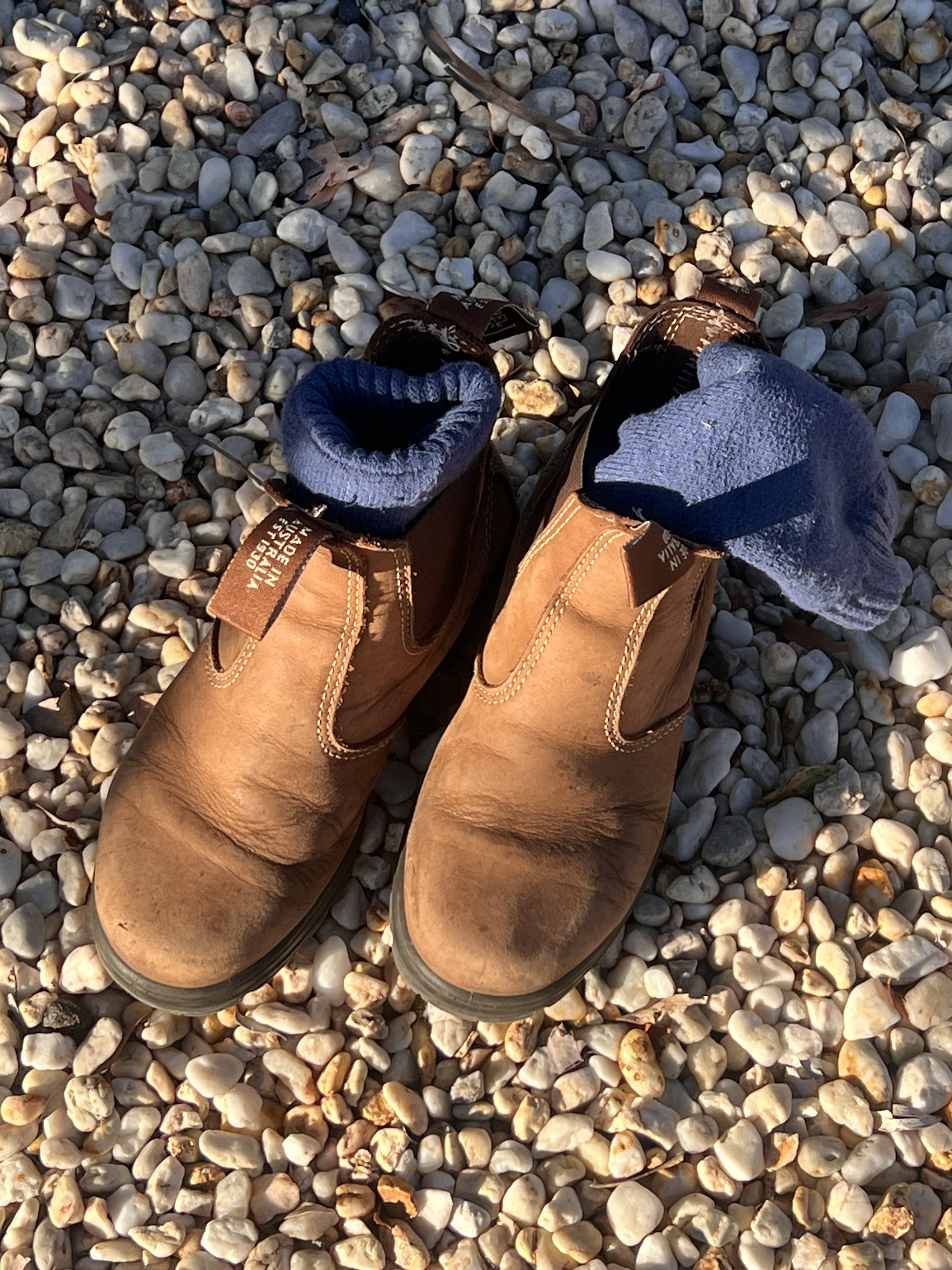 Well-worn brown work boots and thick blue socks resting on pale mine gravel in Queensland gemfields, photographed in warm late afternoon light during Jo and Ella's Rubyvale road trip in search for Sapphires for To Hold & To Have jewellers.