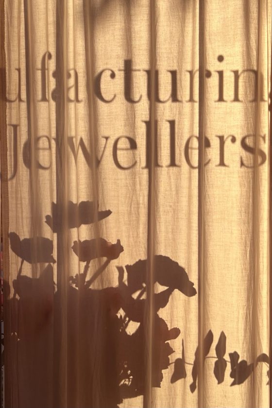 Afternoon sunlight filters through the linen curtain at To Hold & To Have, casting shadows of plants and the words "Manufacturing Jewellers" across the fabric.
